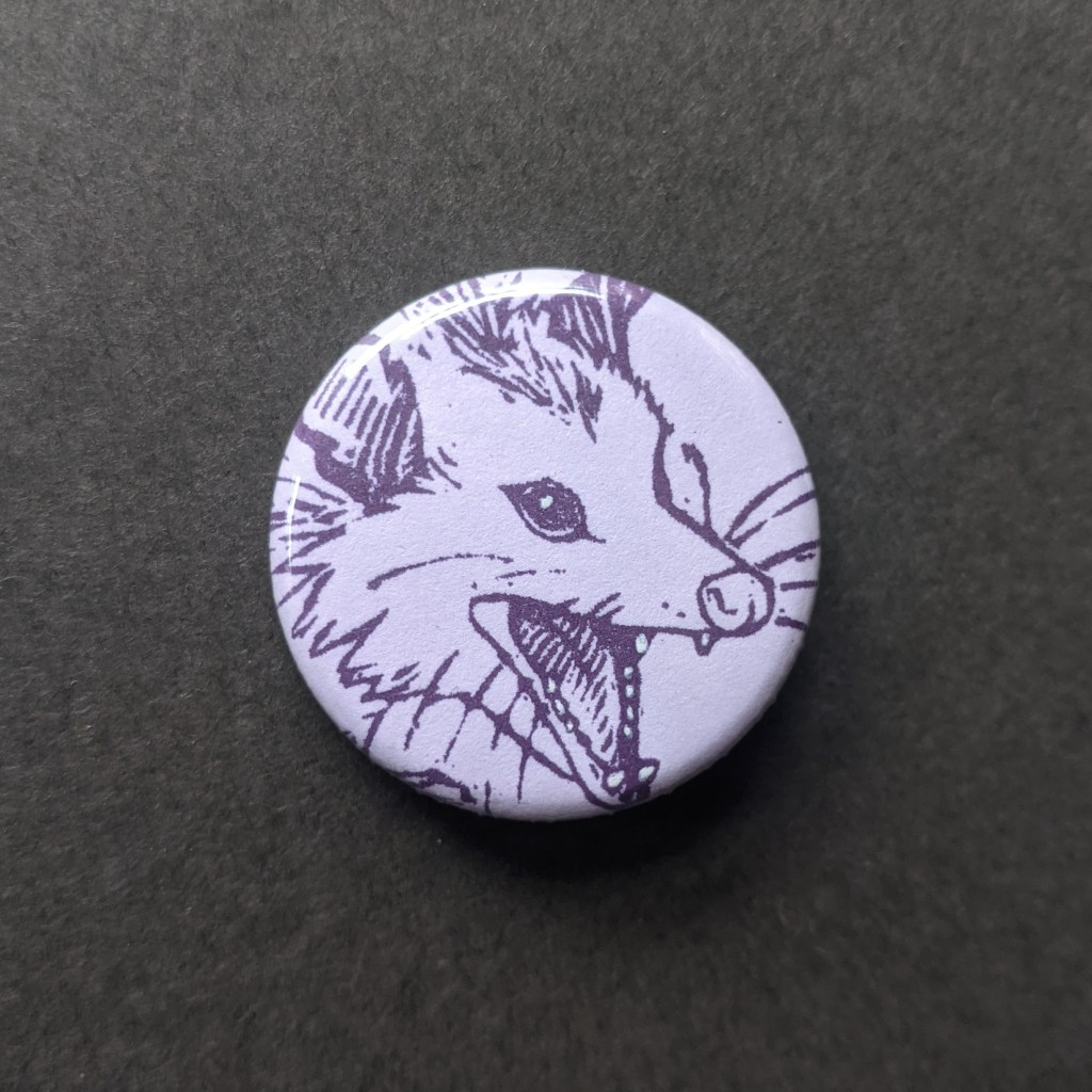 a purple pinback button, stamped with a hand-carved stamp of a opossum