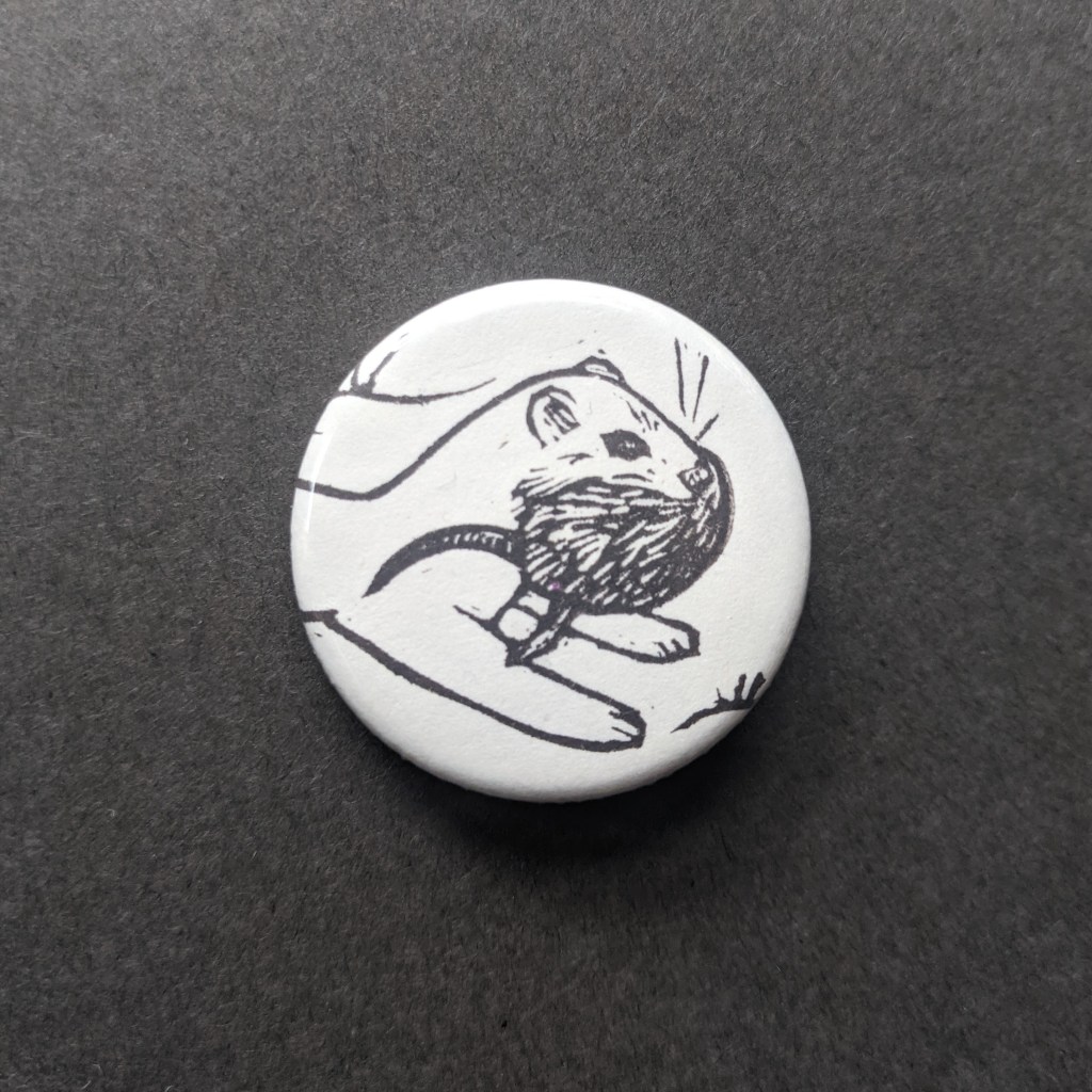 a pinback button, stamped with a hand-carved stamp of an ermine carrying a dead vole in their mouth