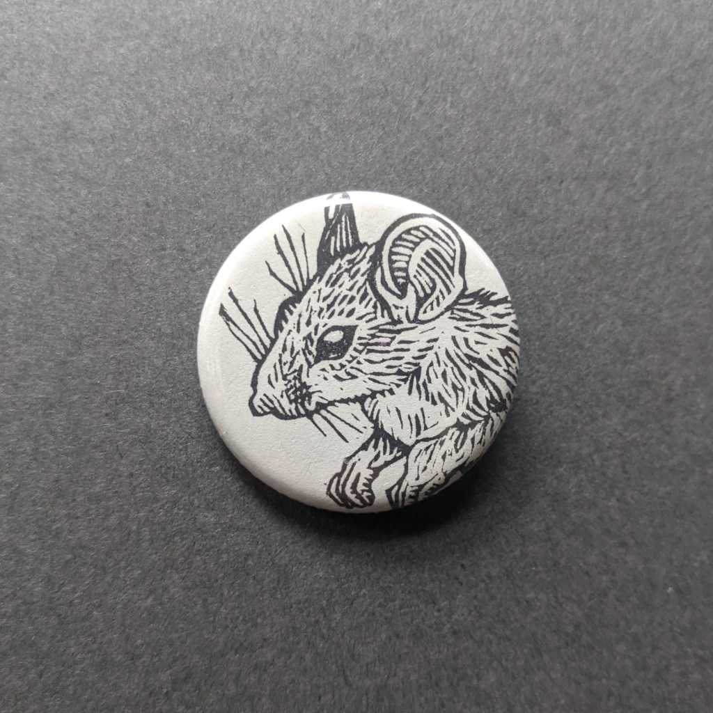 a pinback button, stamped with a hand-carved stamp of a bushy tailed woodrat